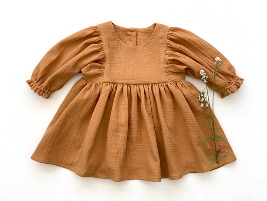 This charming, organic muslin dress is sure to impress! Its caramel-brown hue is perfect for everyday wear and the whimsical style, with its 3/4 length sleeves and ruffled hem with elastic casing, make this knee-length dress a must-have. A wooden button closure in the back completes the look.