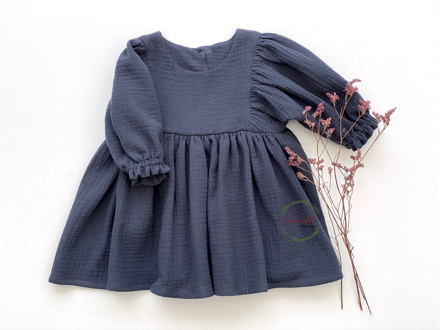 This stylish Denim Blue 3/4 Sleeves Muslin Dress is made from organic muslin and features a whimsical style with ruffled hem and elastic casing. It also has a wooden button closure on the back bodice and reaches the knee length. Perfect for any occasion!