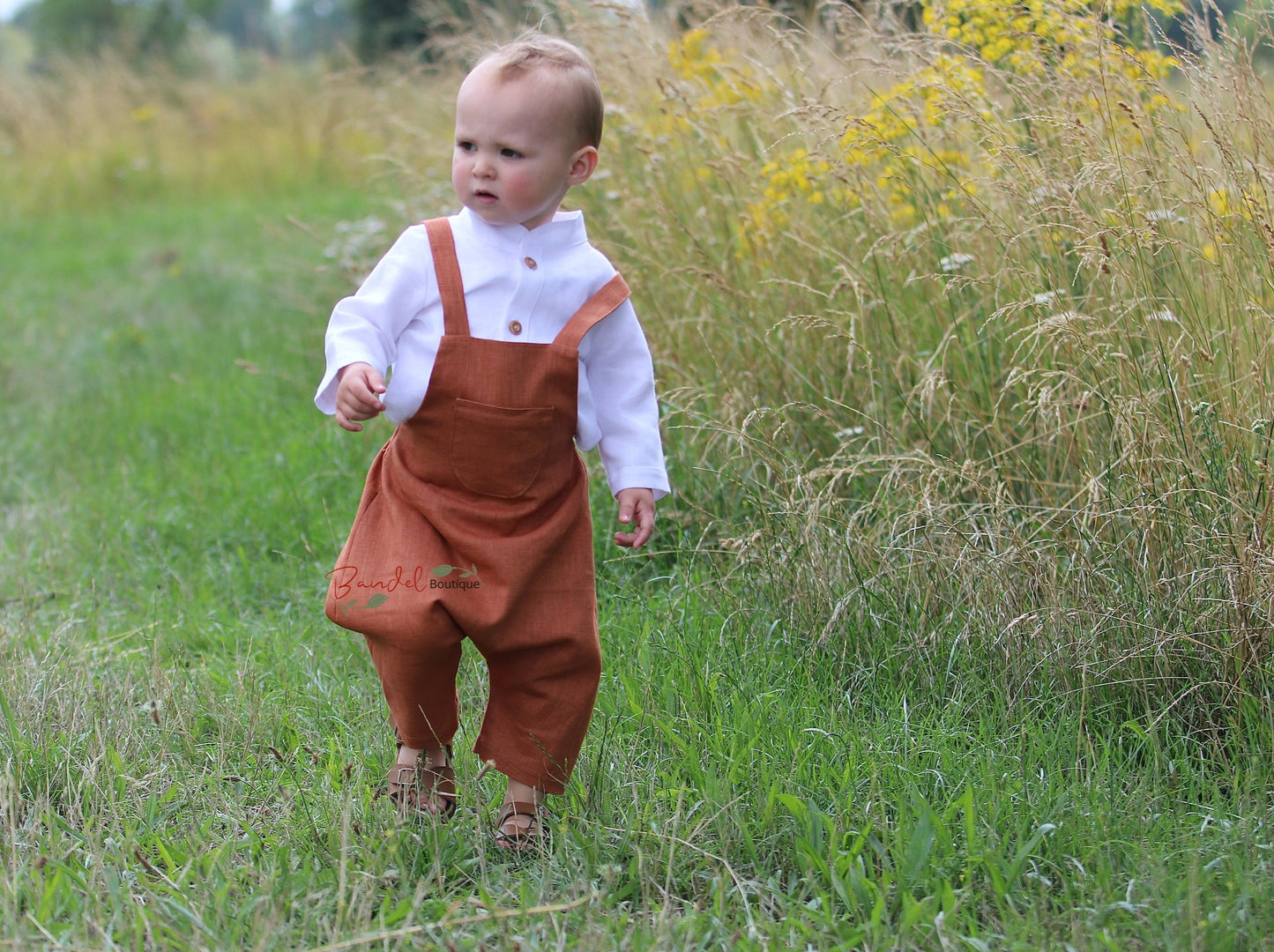 Rust Linen baby romper dungarees feature 3/4 length shorts with a traditional curved front pocket. The elastic back waistband and adjustable tie straps provide a secure fit, and the wooden button closure at the back creates a stylish look.